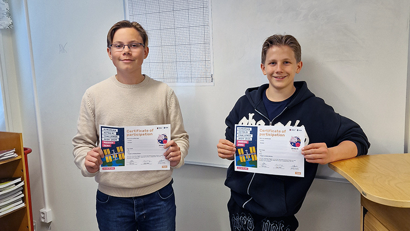 Två pojkar står i ett klassrum och håller upp varsitt diplom.