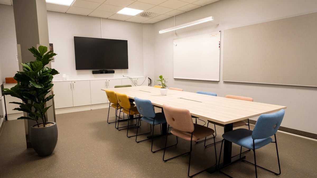 Ett mötesrum med ett avlångt bord omgivet av färgglada stolar, en stor skärm på väggen och gröna växter i rummet.