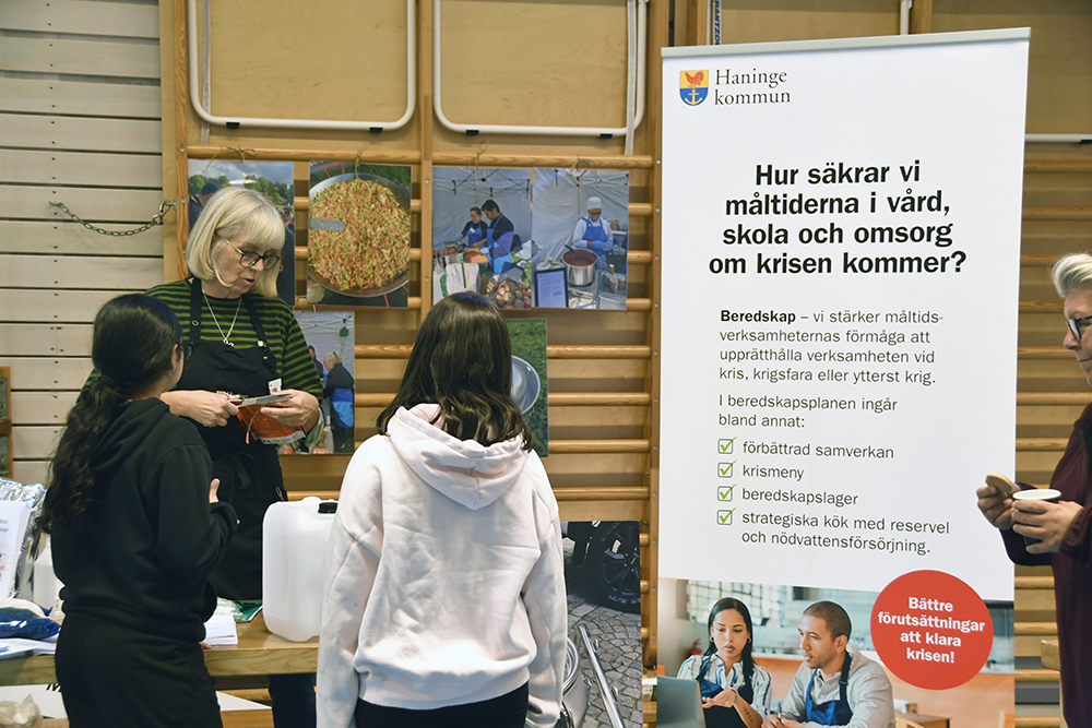 En kvinna, kommunens koststrateg, samtalar med två yngre personer framför en affisch från Haninge kommun om hur måltider säkras inom vård, skola och omsorg vid kris.