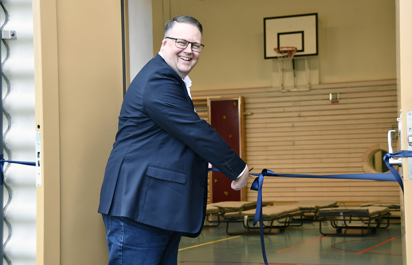 Jens Davidson (KD), kommunalråd i Haninge kommun, klipper ett blått band vid invigningen av trygghetspunkterna. I bakgrunden syns en gymnastiksal med uppställda fältsängar och en basketkorg.