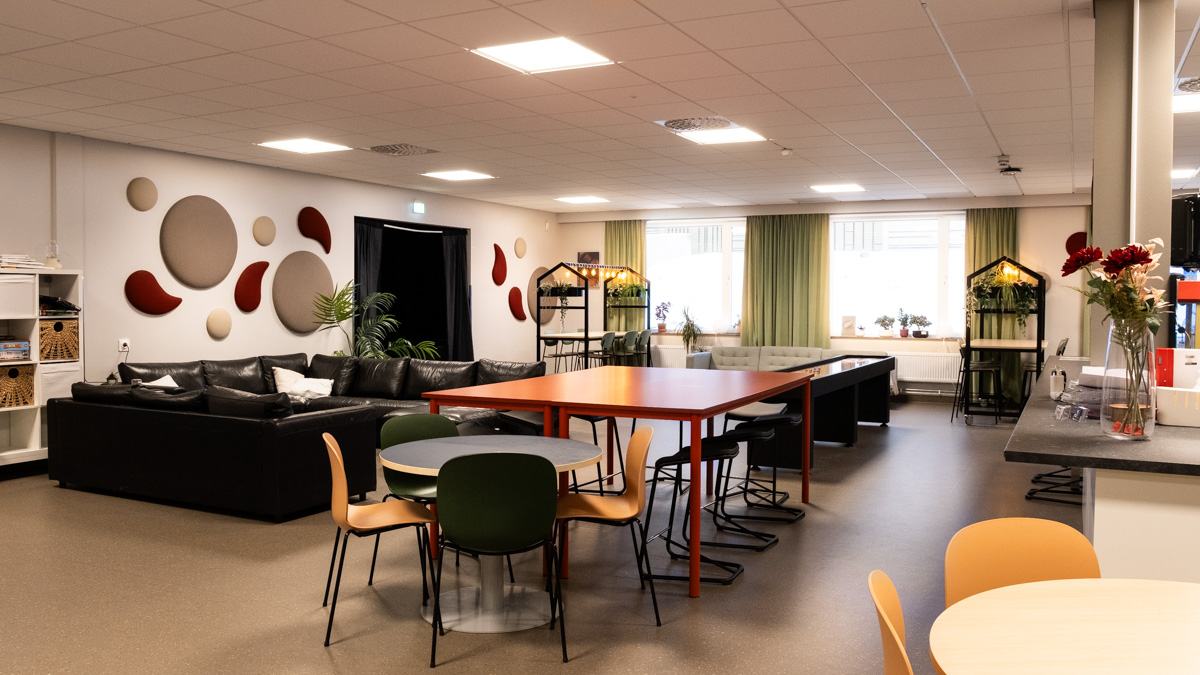 Gemensam lounge med svart soffa längs väggen, flera bord och stolar i olika färger i förgrunden samt ett större rött bord i mitten. 