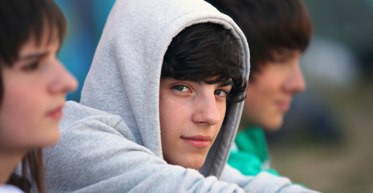 Tre tonåringar sitter bredvid varandra och en pojke med luvan på ser in i kameran. 